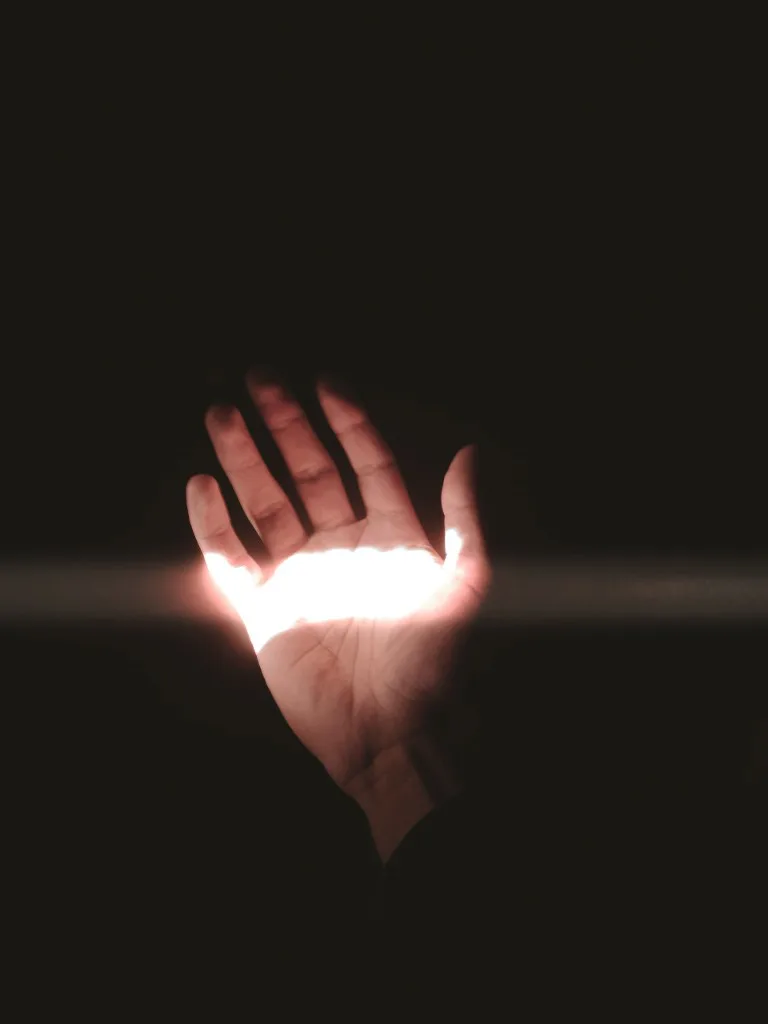 An open hand reaching upward with a beam of light passing through the palm against a dark background