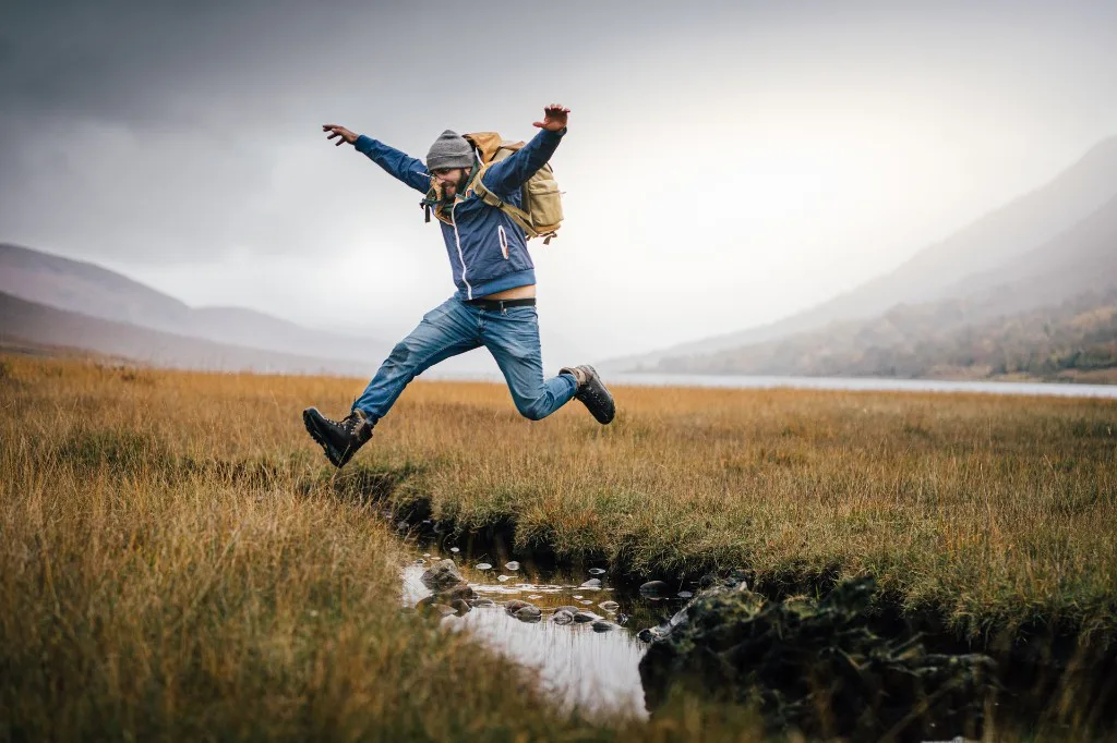 A hiker jumping across a rocky stream in an open mountain landscape